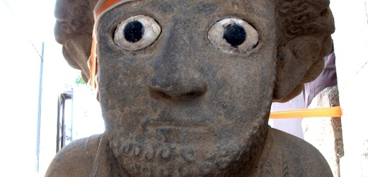 Kudrnaté kadeře připomínají helmu, velké oči sochař vytvořil vsazením barevných kamenů.
