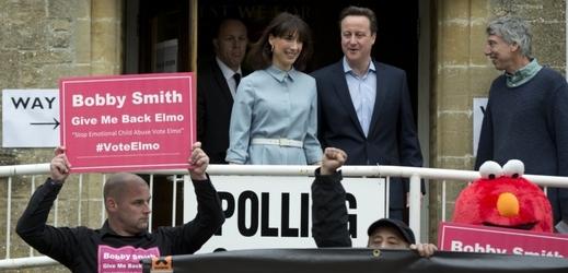 David Cameron s manželkou vycházejí z volební místnosti, která je obložená protestujícími.