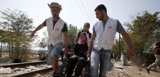 Humanitární pracovníci nesou postiženého migranta na invalidním vozíku, hranice Řecka a Makedonie.