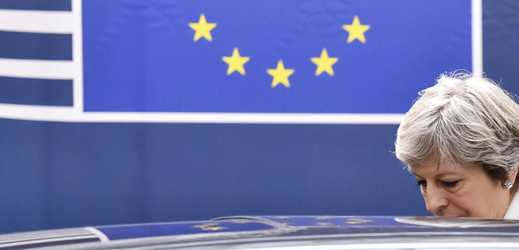 Theresa Mayová nastupuje do auta. V pozadí vlajka EU. 