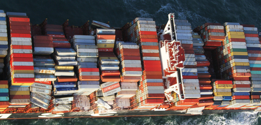 Letecký snímek nákladu lodi, na kterém jsou vidět popadané řady kovových kontejnerů.